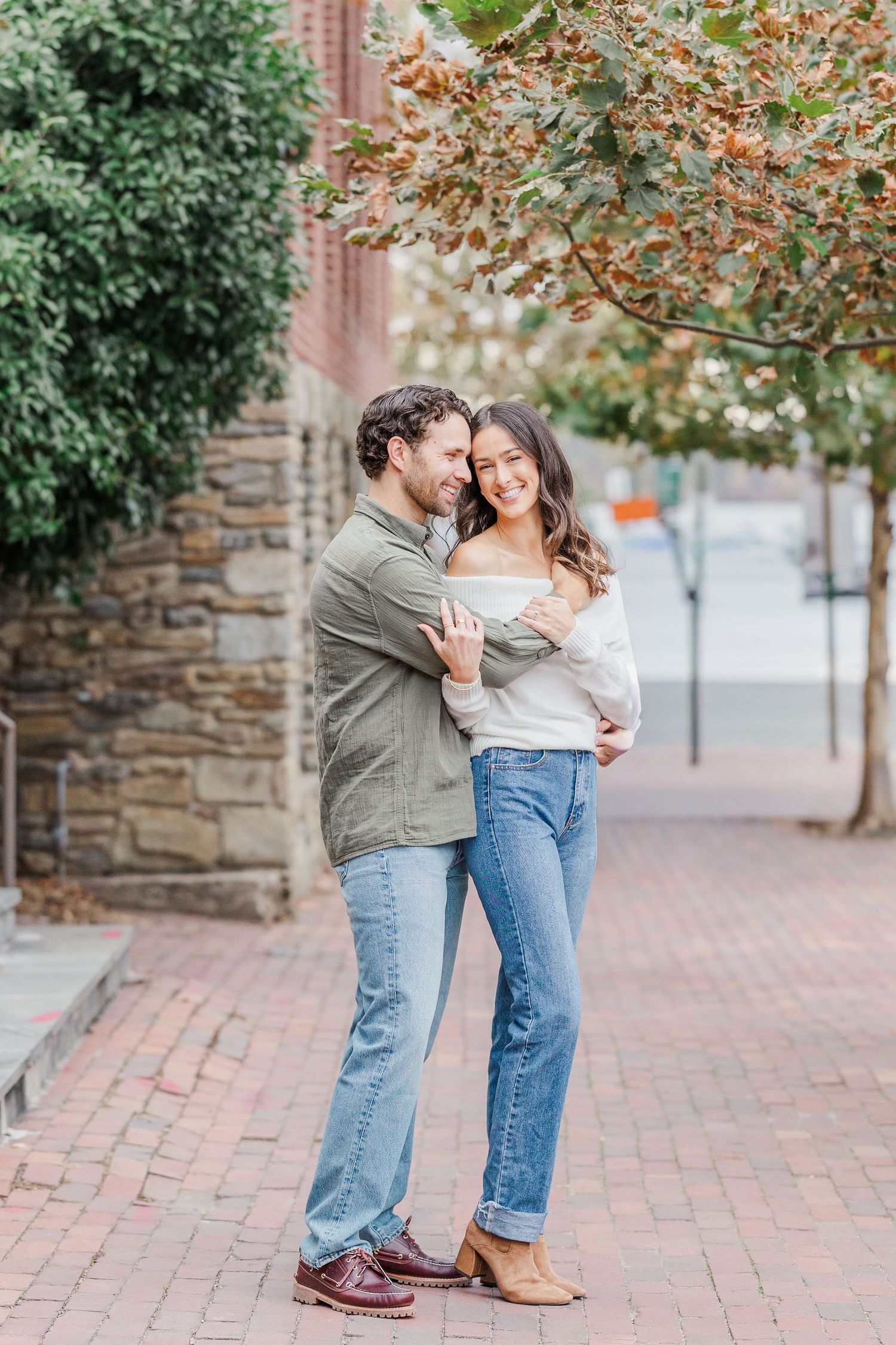 A romantic couple shares joyful moments on a brick sidewalk near a stone wall during an autumn engagement photoshoot.