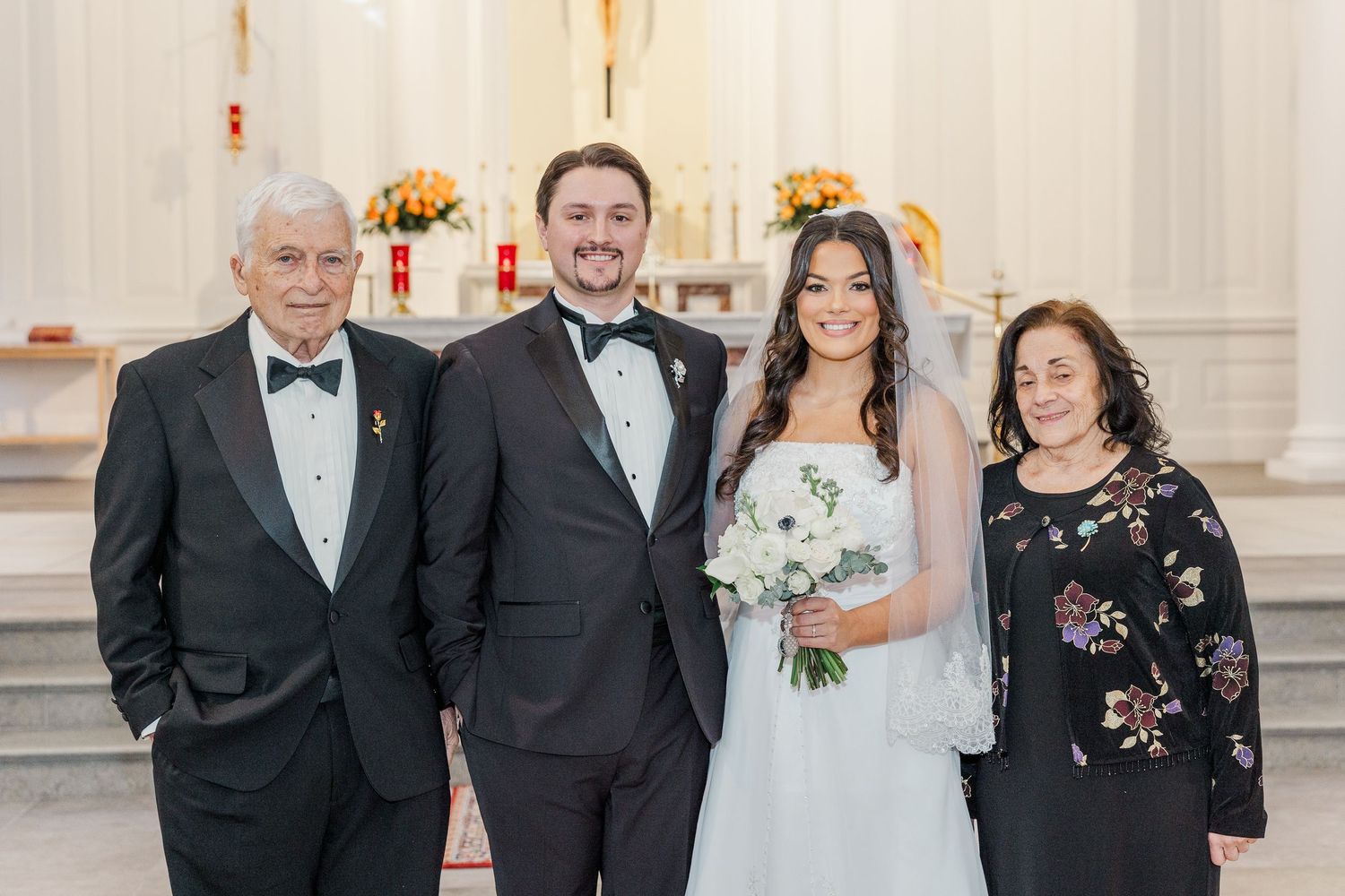 A group of formally dressed people pose together at an elegant wedding venue with white floral arrangements.