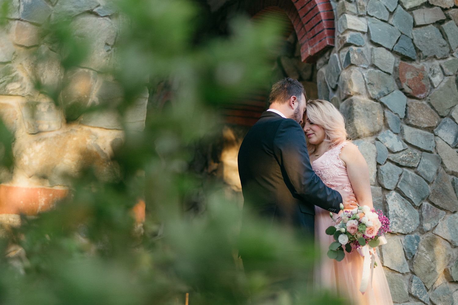 A couple embraces during their wedding ceremony at a stone wall venue with soft natural lighting and greenery.