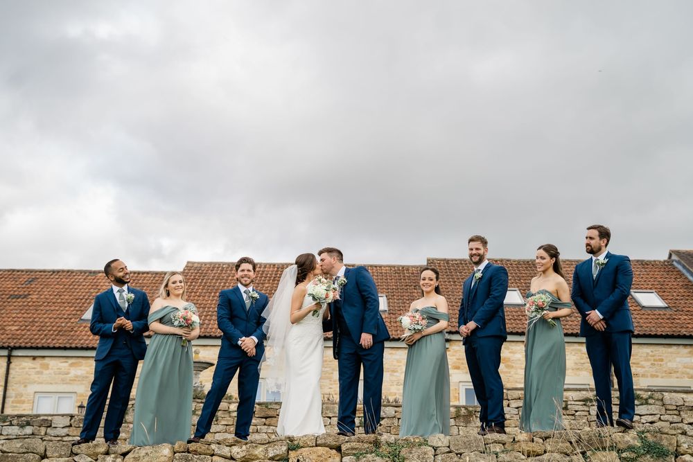 Wedding party in formal attire poses playfully in a line against cloudy sky and brick building backdrop.