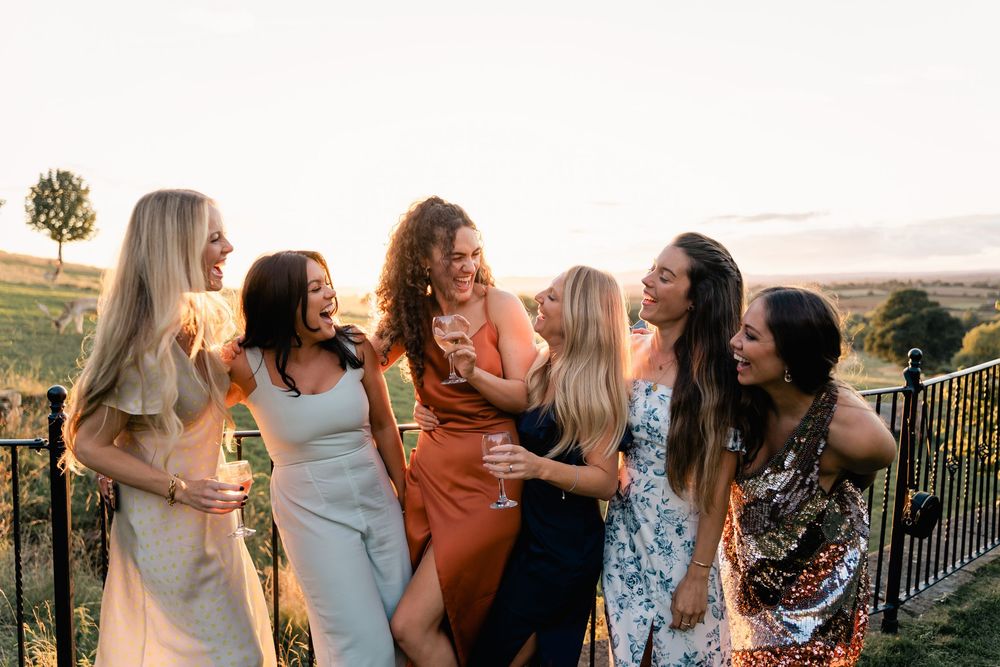 A group of friends laugh and embrace while enjoying sunset views from a balcony overlooking mountains.