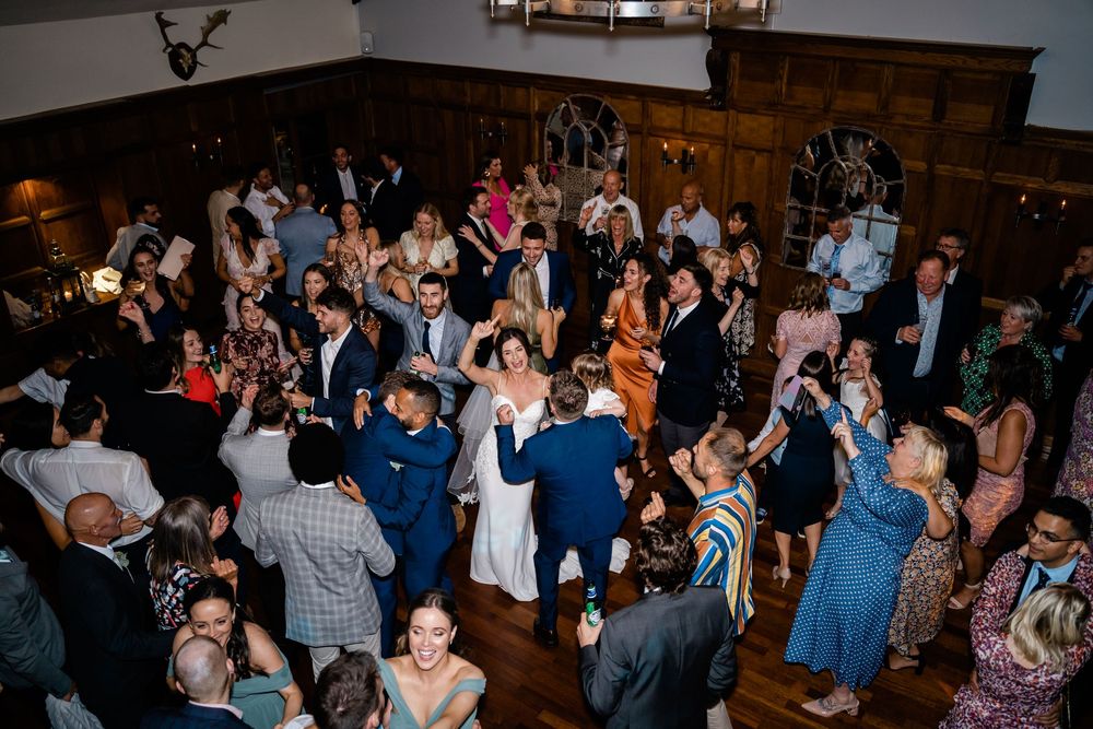 Wedding reception guests dance and celebrate together on a rustic wooden dance floor at an indoor venue.