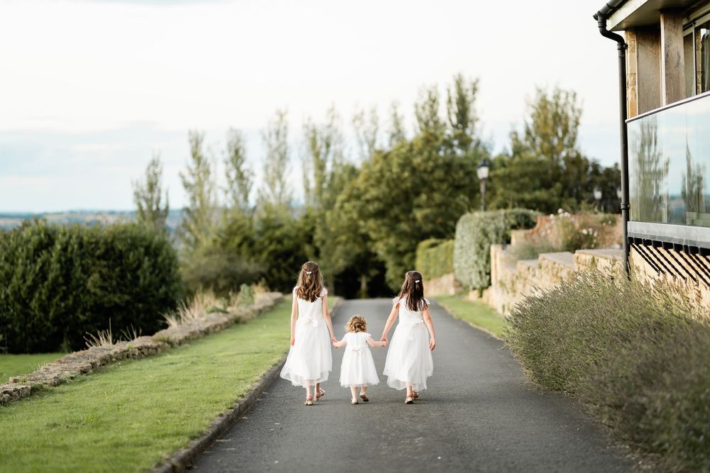 Three people in flowing white dresses walk along a scenic countryside path at sunset.