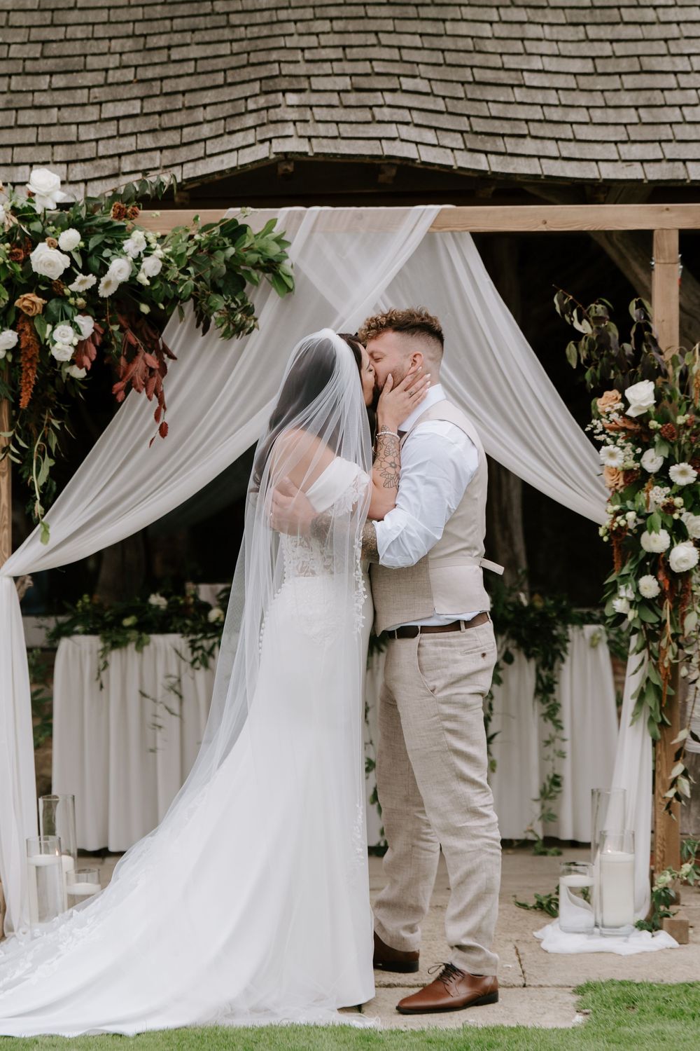 A romantic wedding ceremony sequence showing a couple sharing their first kiss under white draped fabric and floral decor.