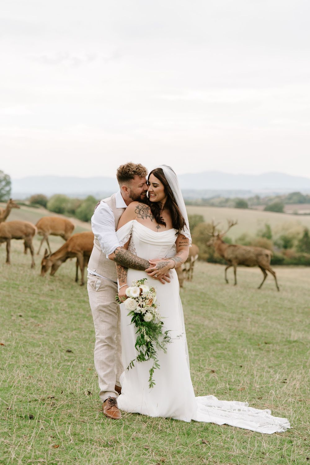 A romantic wedding couple embraces in a scenic field with horses grazing in the background on a sunny day.
