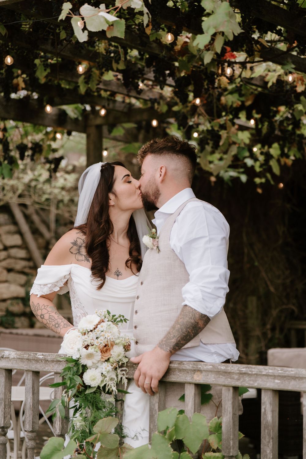 A romantic wedding moment captured on a rustic wooden bridge surrounded by greenery and delicate white florals.