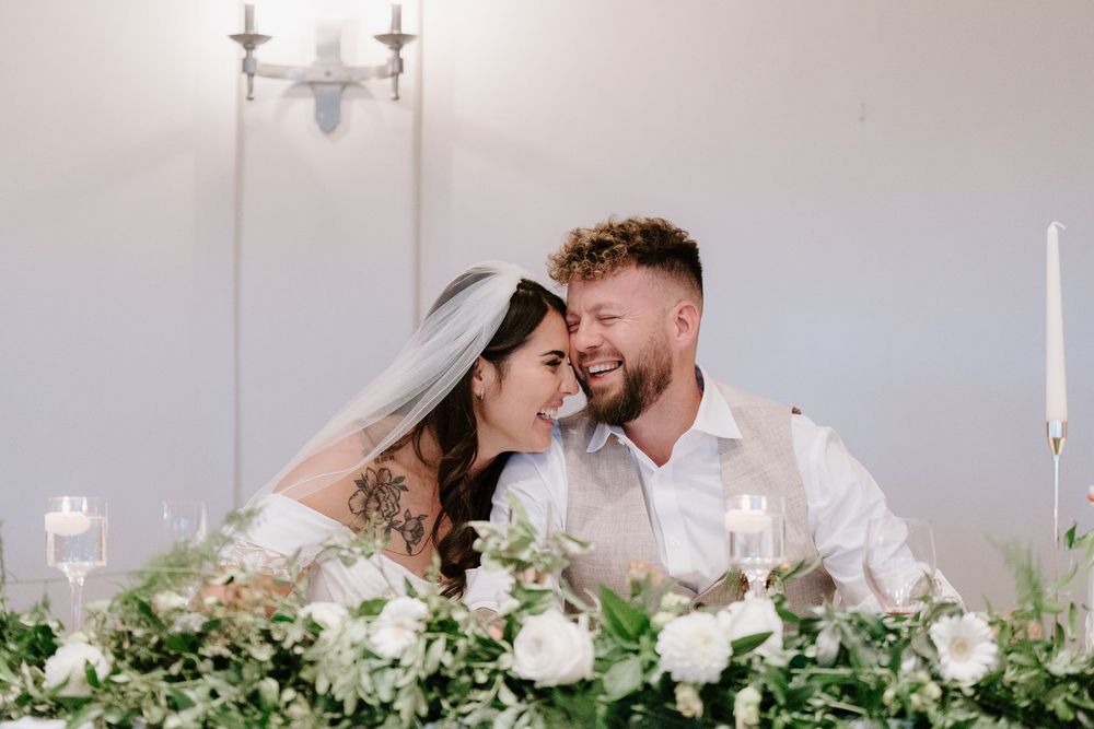 A romantic wedding couple shares an intimate moment behind a lush floral arrangement at their reception table.