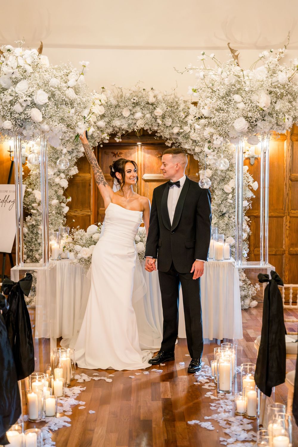 A romantic indoor wedding ceremony setup with white floral arches, glowing candles, and a wooden floor aisle.