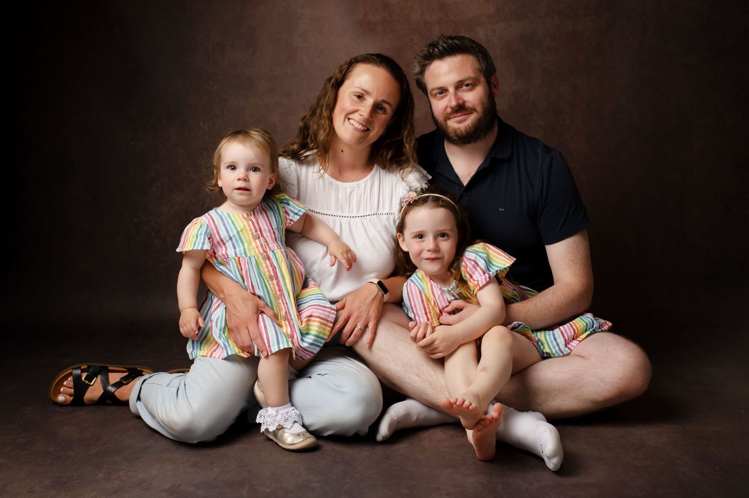 A family of four wearing coordinated white and beige outfits poses together on a wooden bench with dried pampas grass decor.