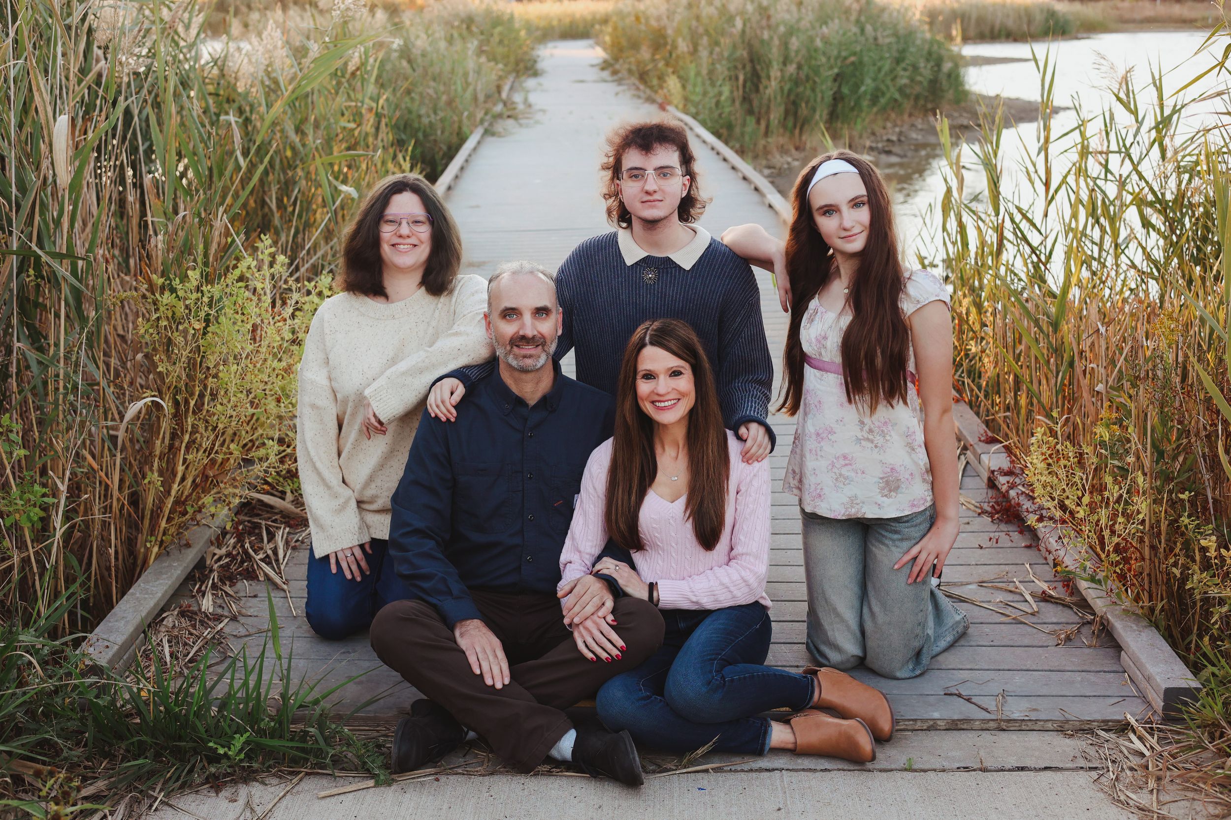 A family group poses together on a wooden boardwalk surrounded by tall grass.