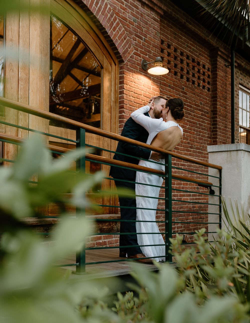 A couple shares a romantic moment on an outdoor balcony against a rustic brick wall at dusk.