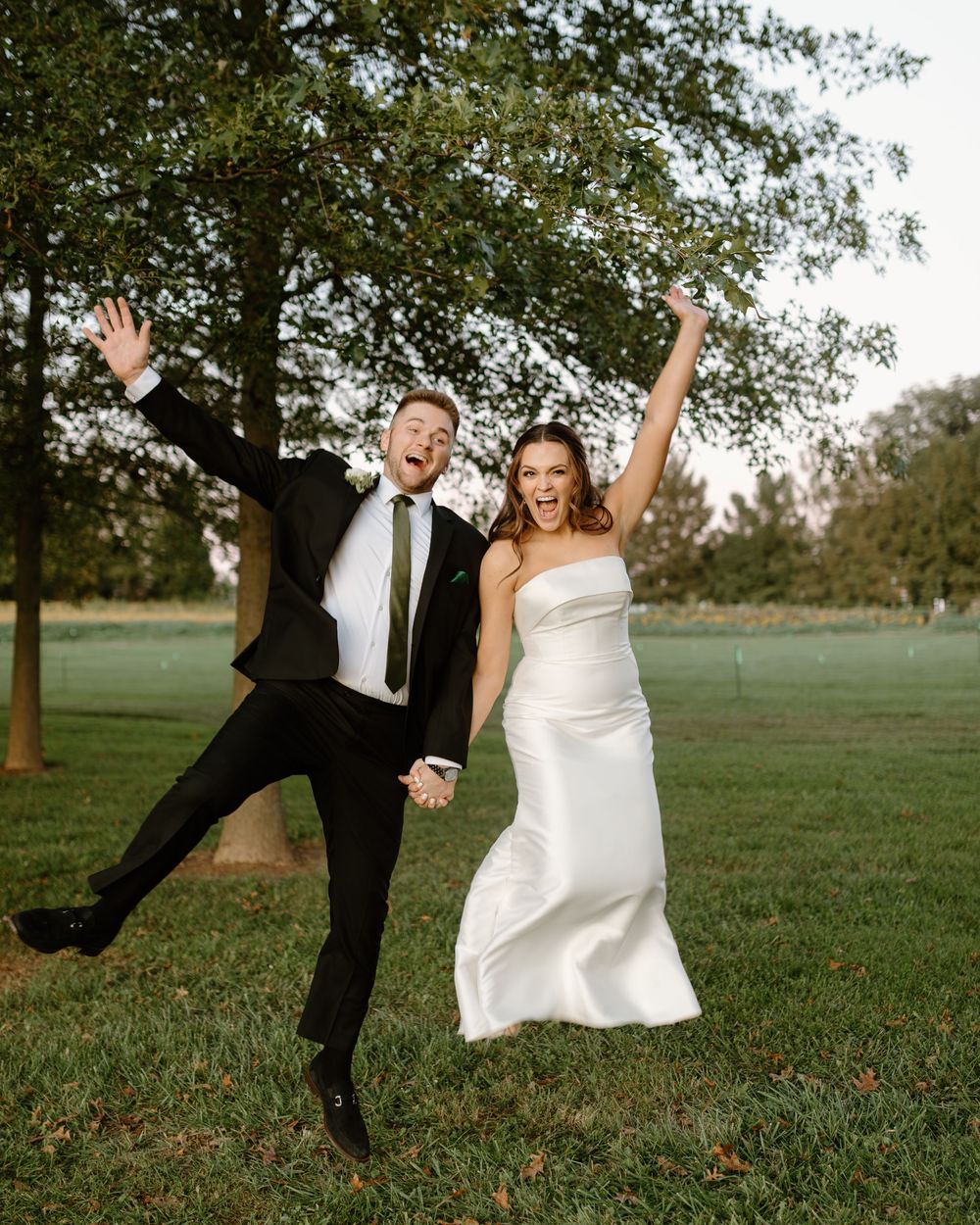 A couple in wedding attire playfully jumps and poses together on a grassy field during their outdoor photo session.
