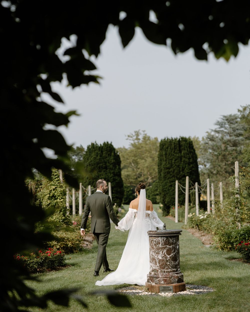 A romantic garden wedding scene with a couple walking through manicured hedges and decorative stone columns.