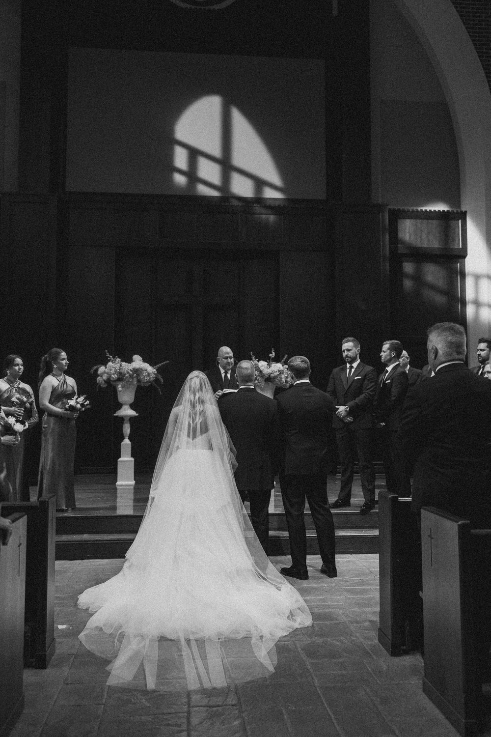 A bride in a flowing white gown and veil stands at the altar during an indoor church wedding ceremony.
