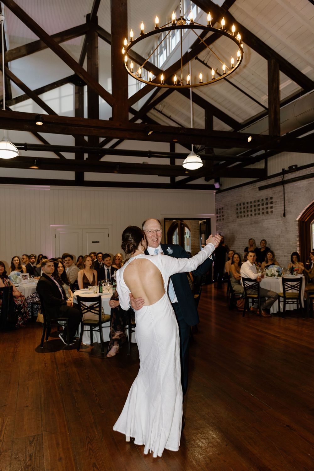 A wedding dance takes place on a wooden floor under a rustic beamed ceiling with a circular chandelier.
