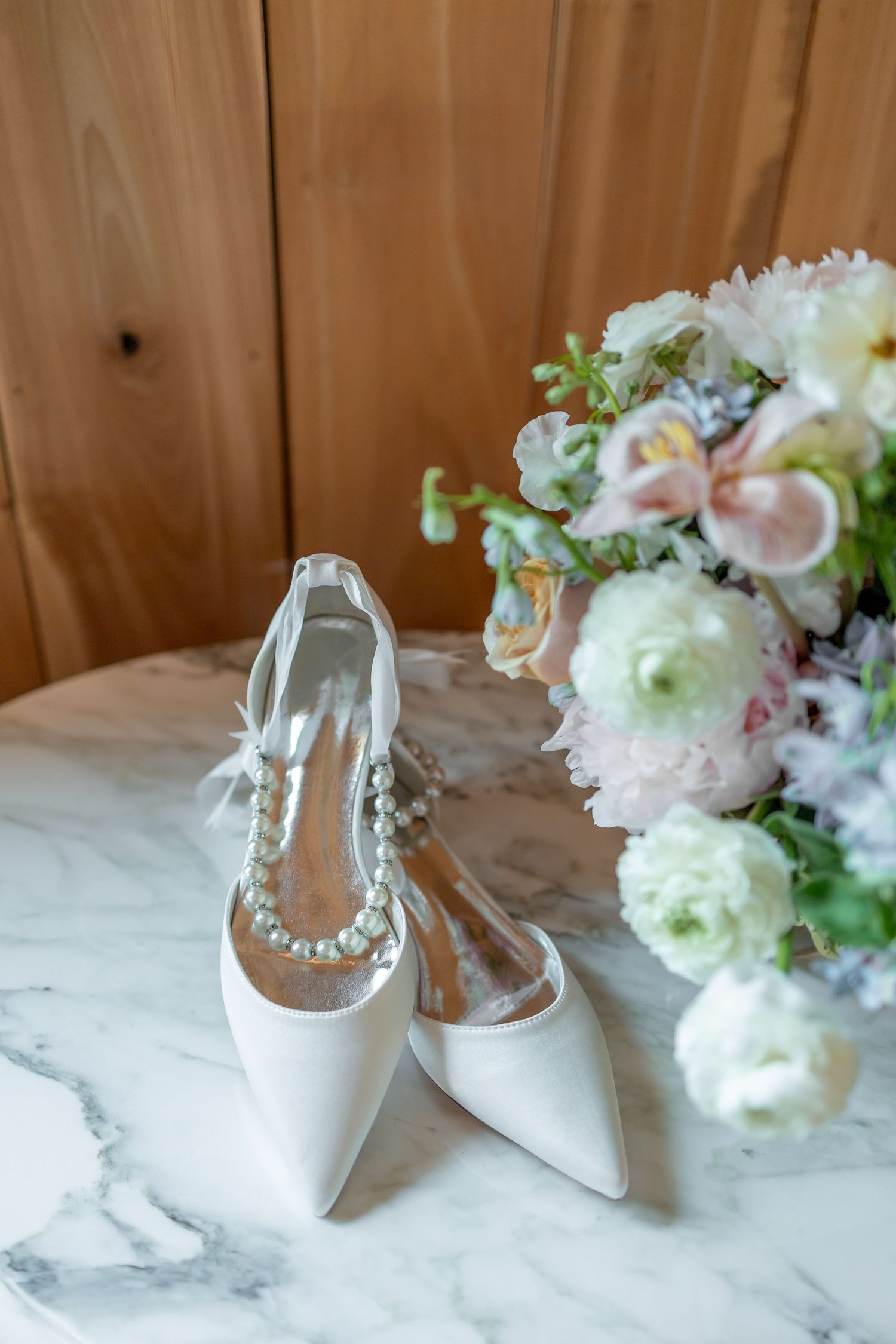 Elegant white bridal shoes with pearl details displayed alongside delicate floral arrangements on a marble surface.