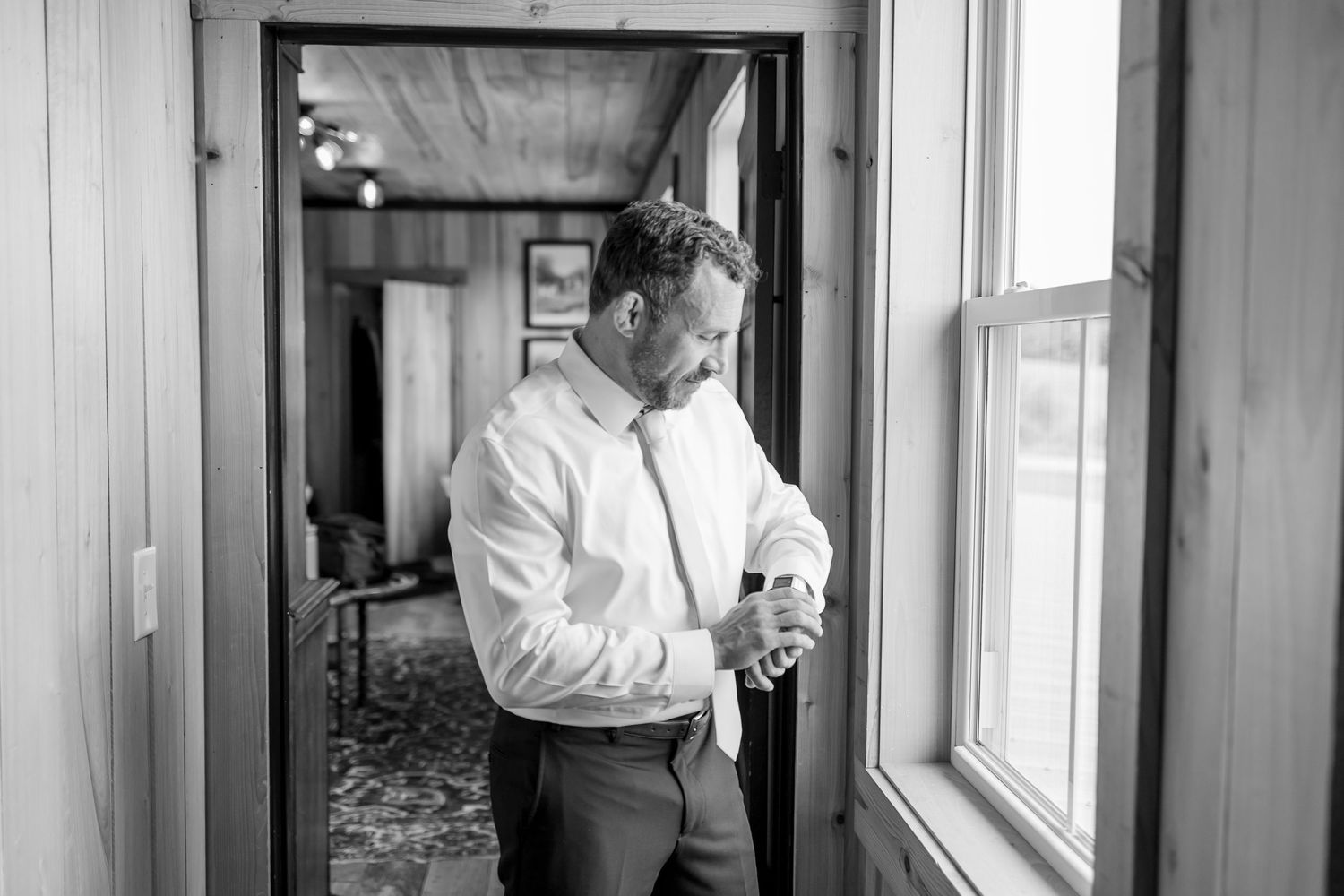 A black and white photo of a person looking out a window in a rustic interior setting.