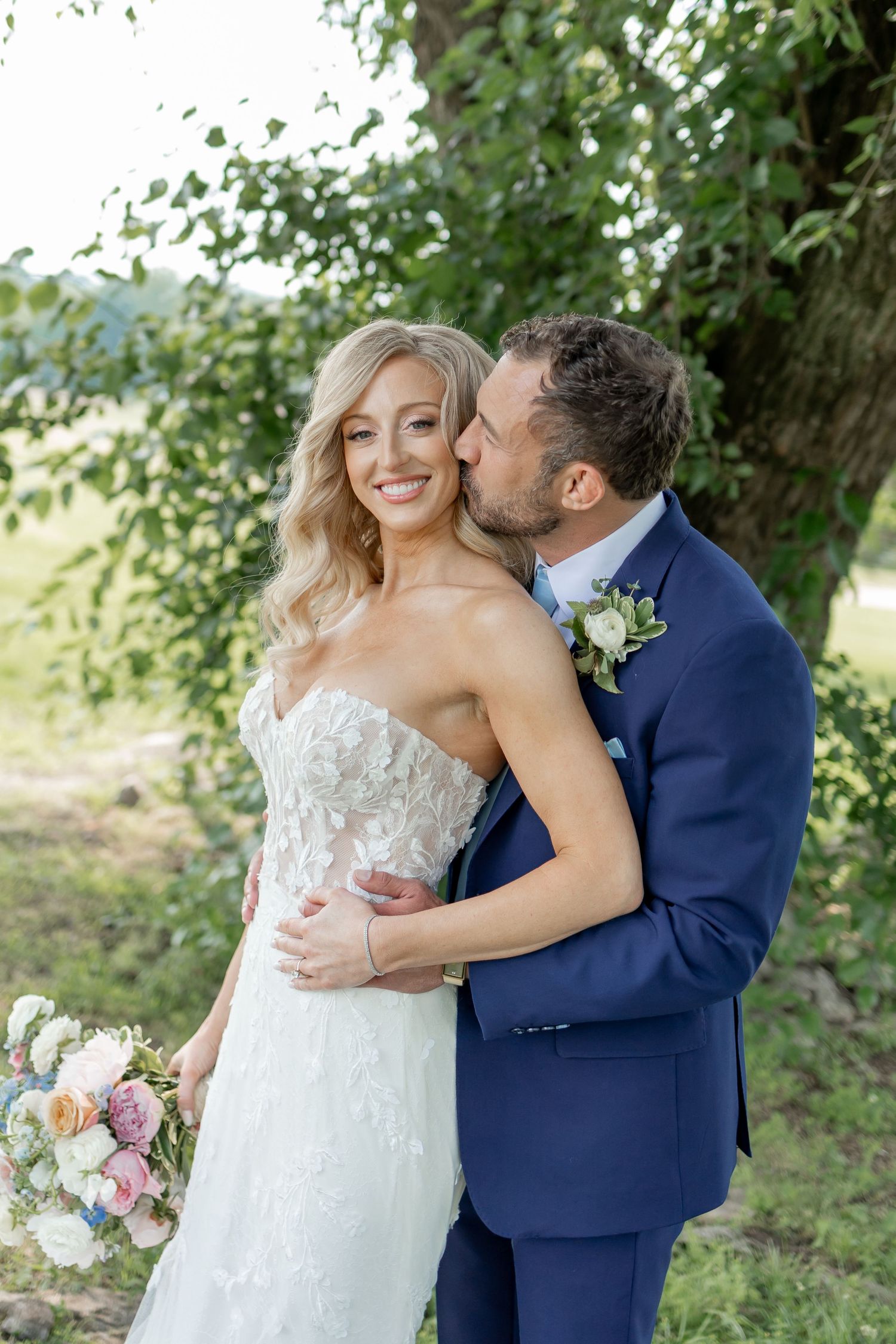 A romantic wedding moment captured outdoors with the bride in white lace and groom in navy suit among green foliage.