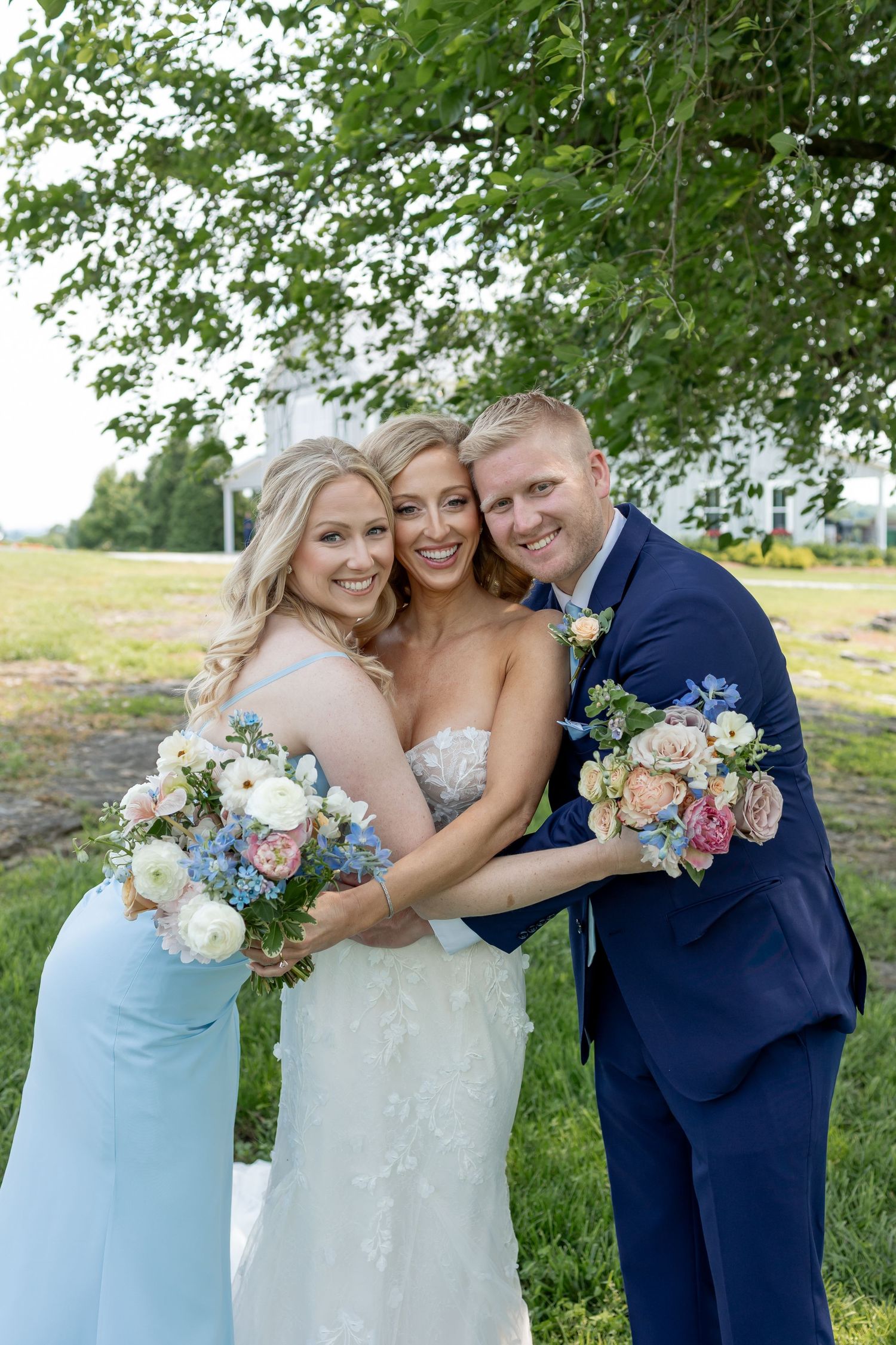Wedding party poses outdoors with light blue bridesmaid dresses and navy suit against green foliage background.