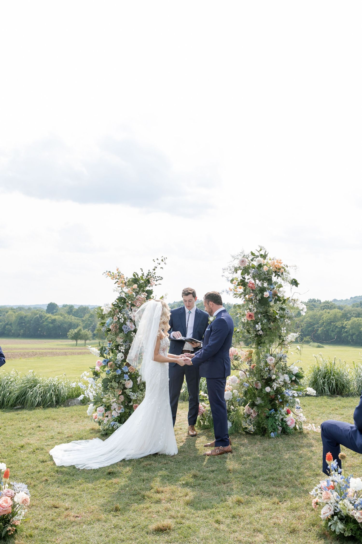 Outdoor wedding ceremony under floral arch with white and pink roses in countryside setting with rolling hills.