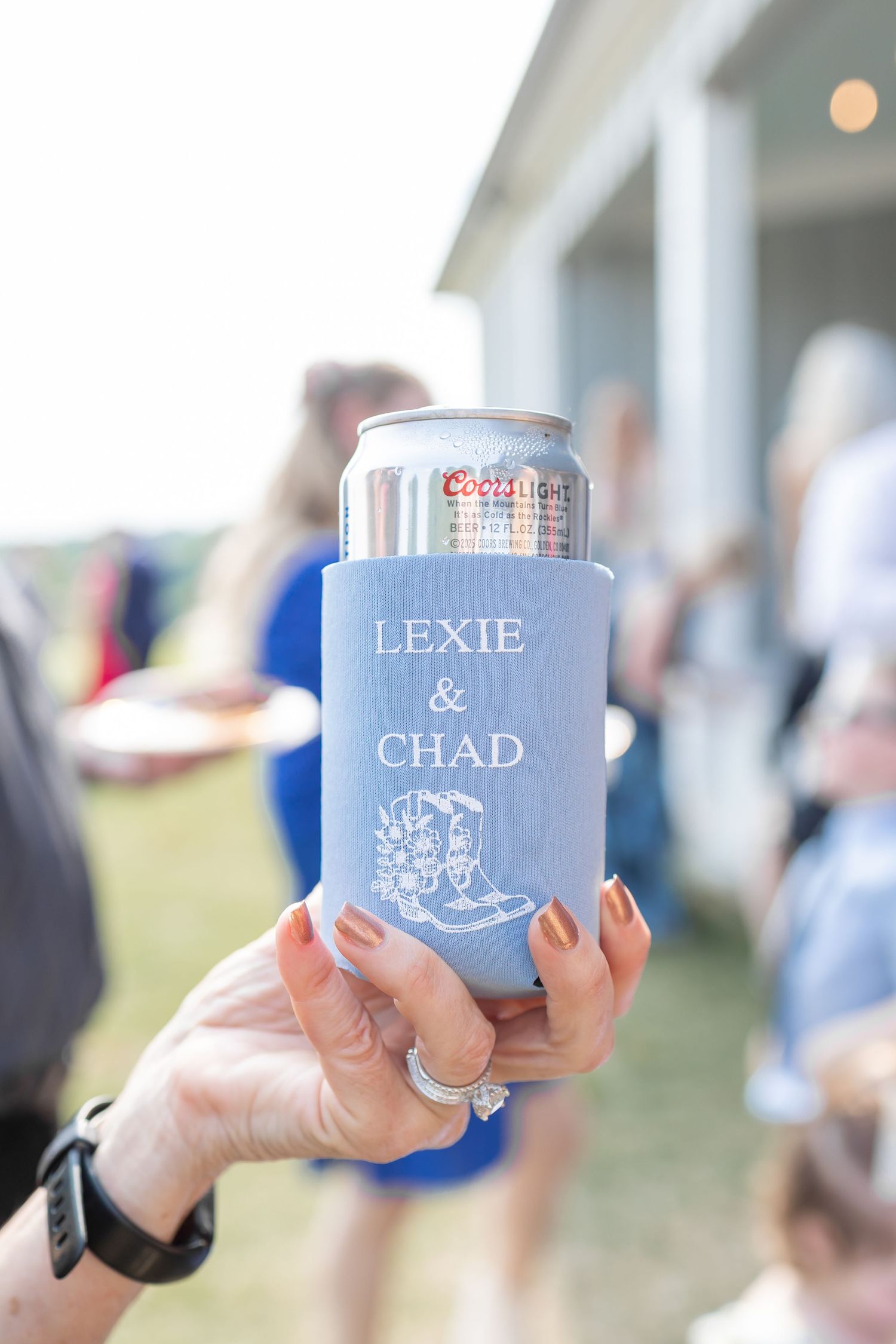A series of hands holding personalized blue and white drink koozies at an outdoor event.