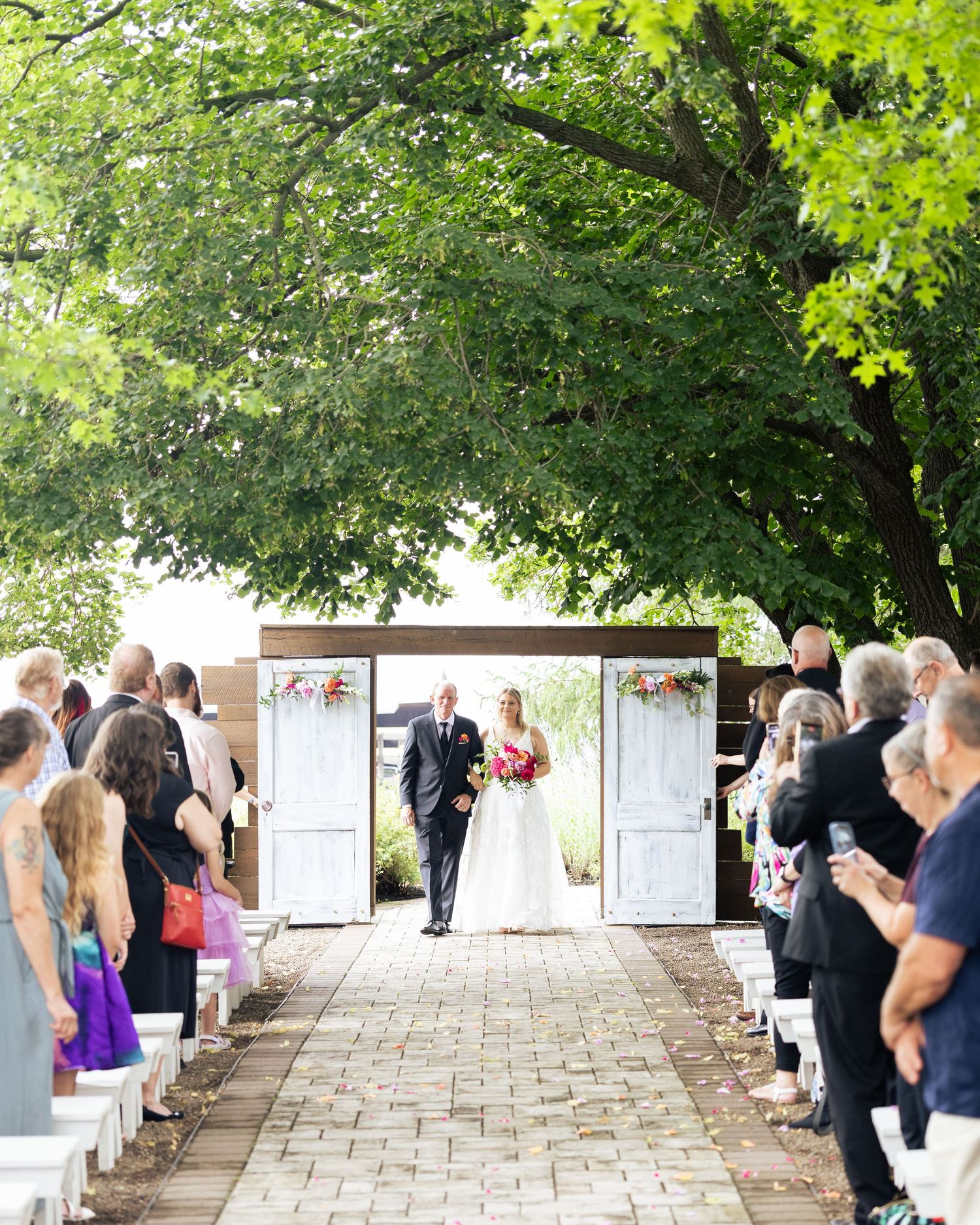 Bride in white gown walks down outdoor aisle at sunny woodland wedding ceremony, holding bouquet.