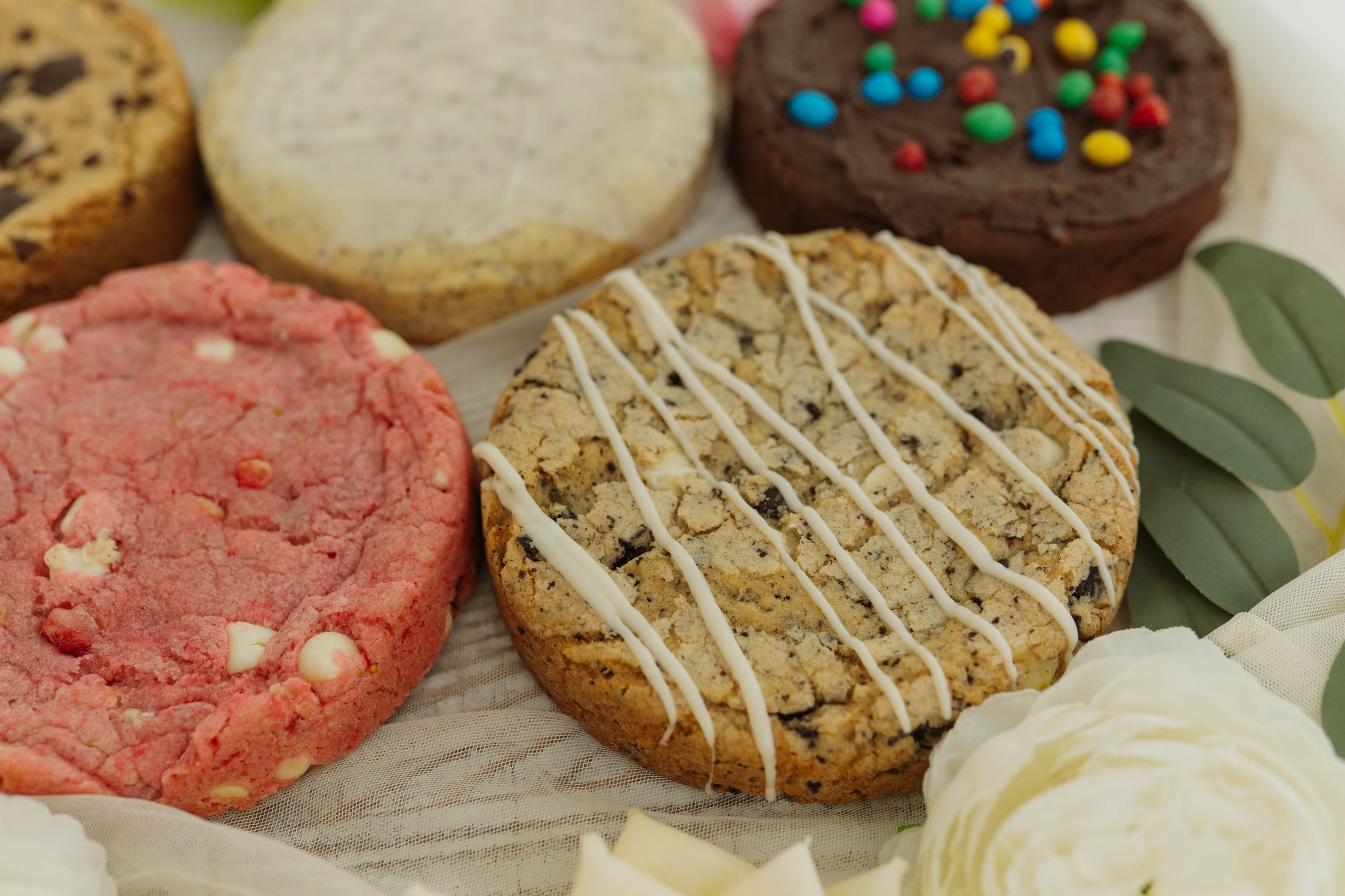 Colorful homemade cookies with sprinkles and drizzles displayed among fresh flowers.