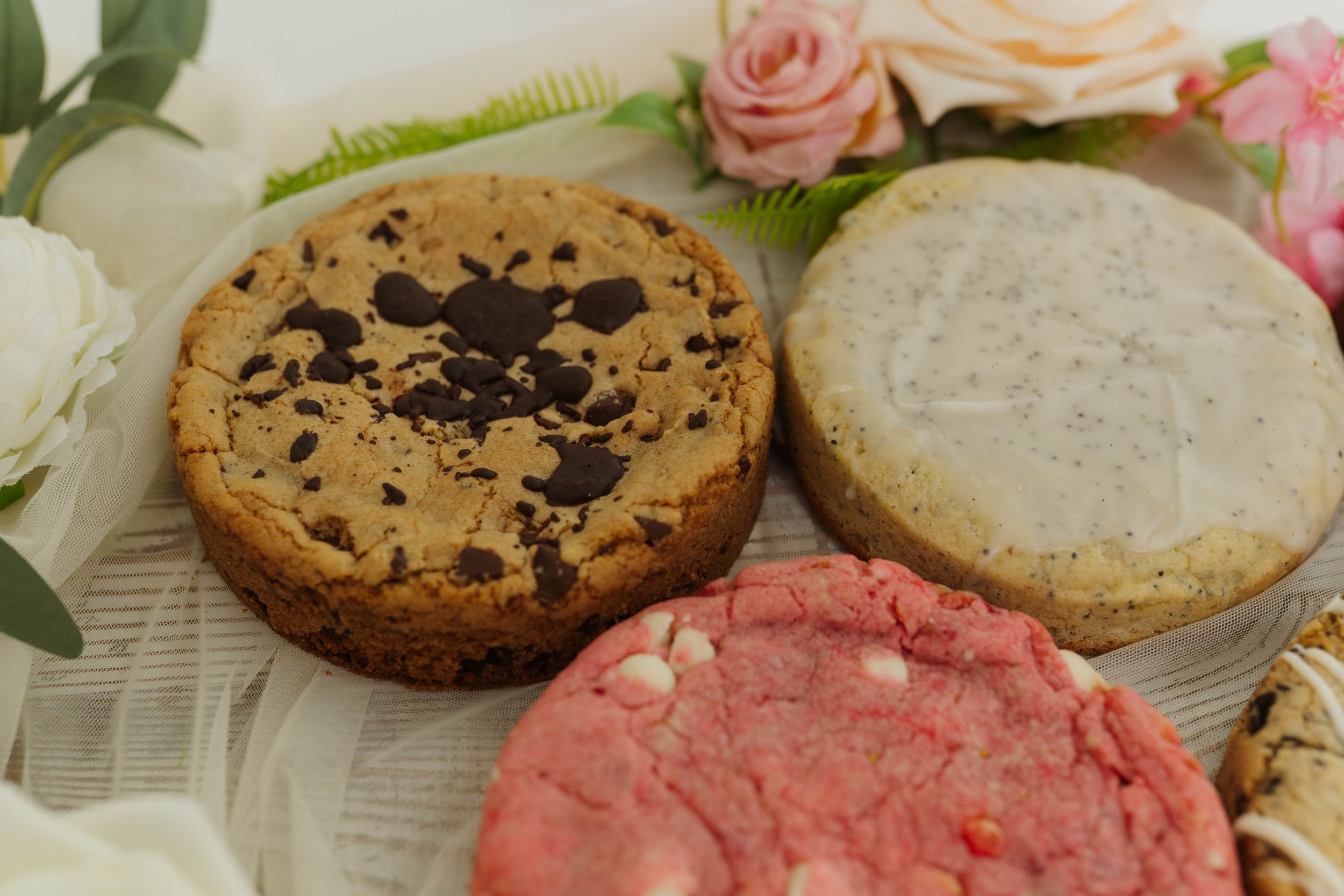 Gourmet cookies with chocolate chips and pink frosting displayed with white roses.
