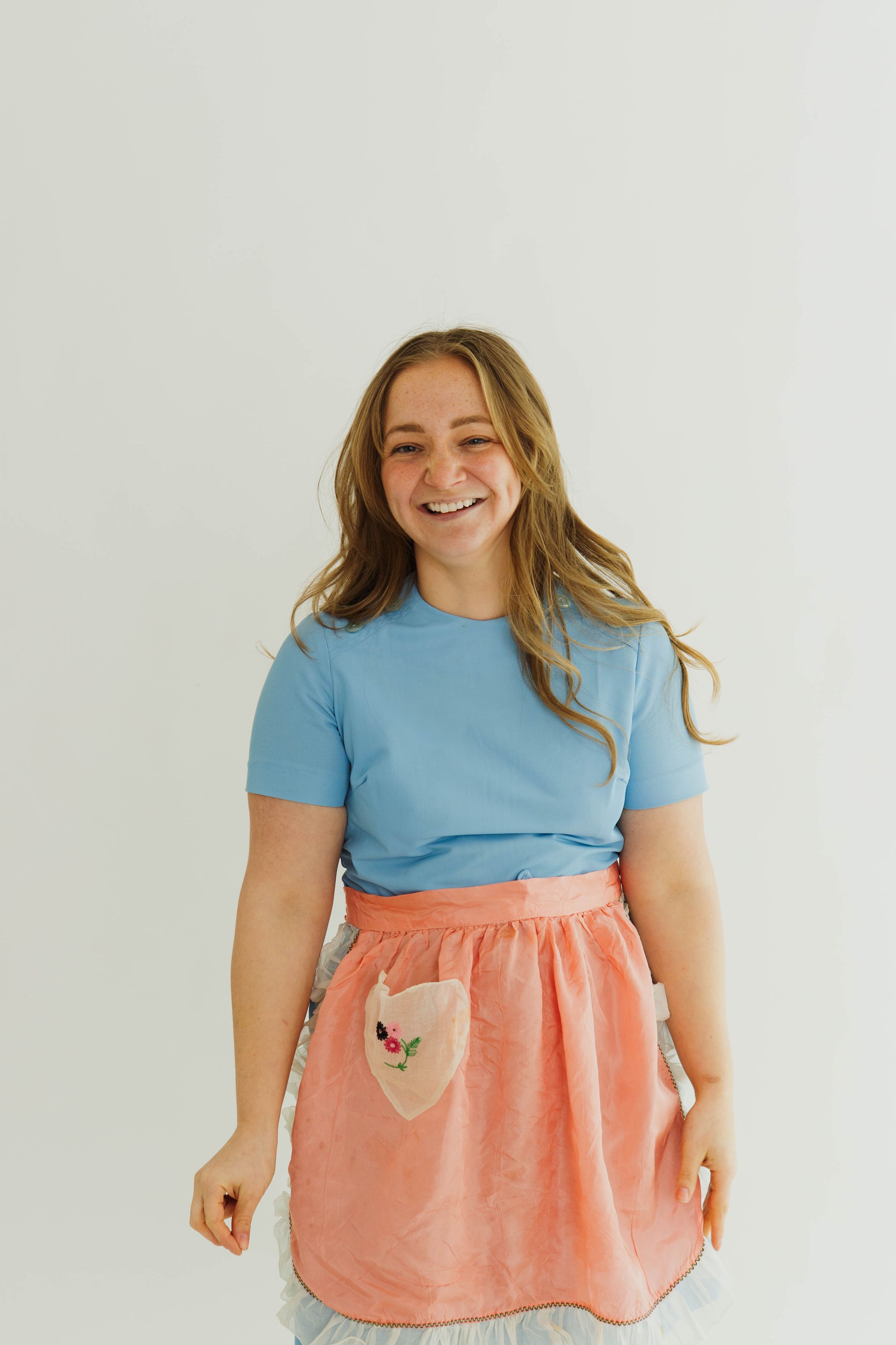 Person wearing blue t-shirt and coral skirt with pocket detail poses against white wall.