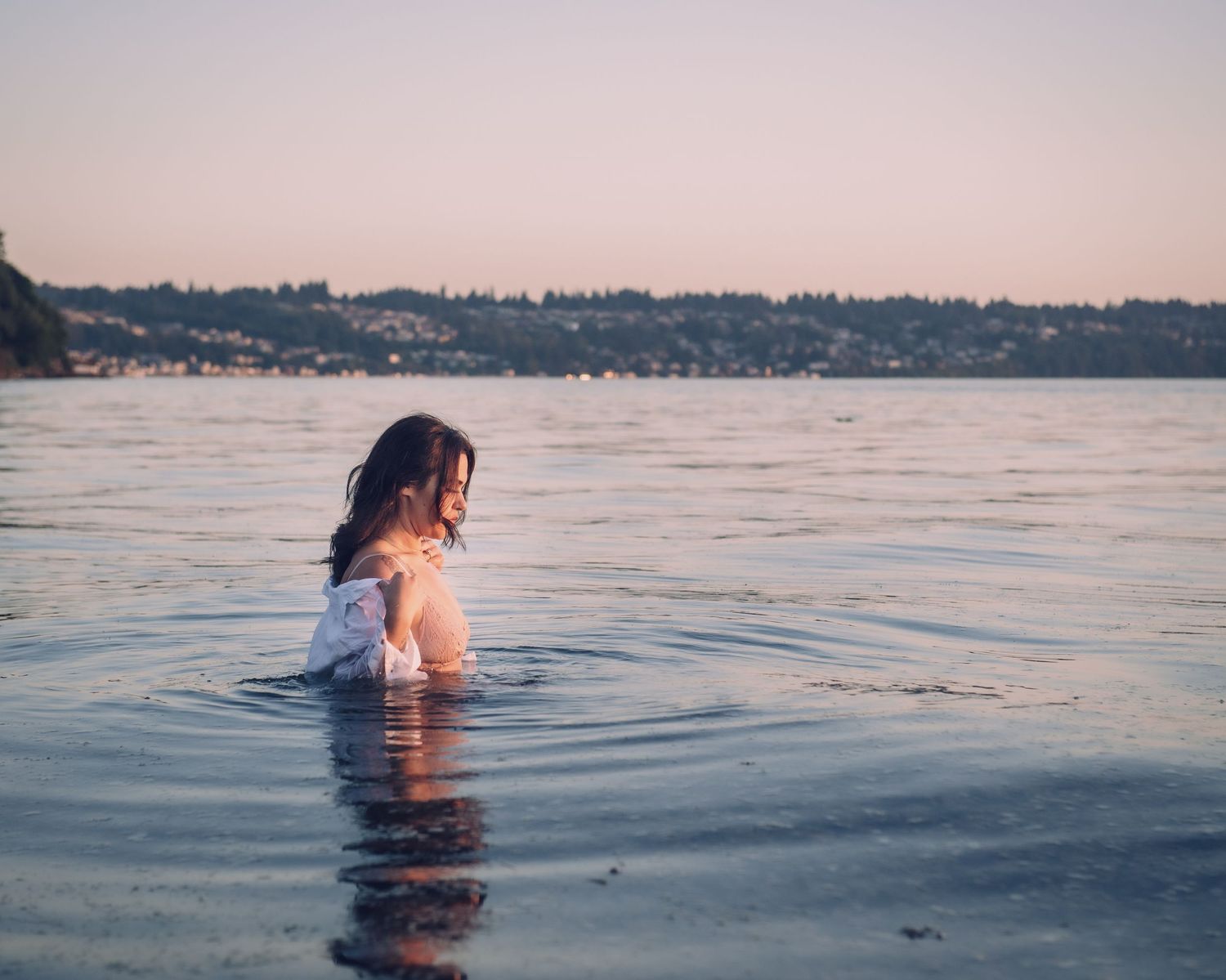 Person wading in calm lake waters at dusk with tree-lined shore in background.
