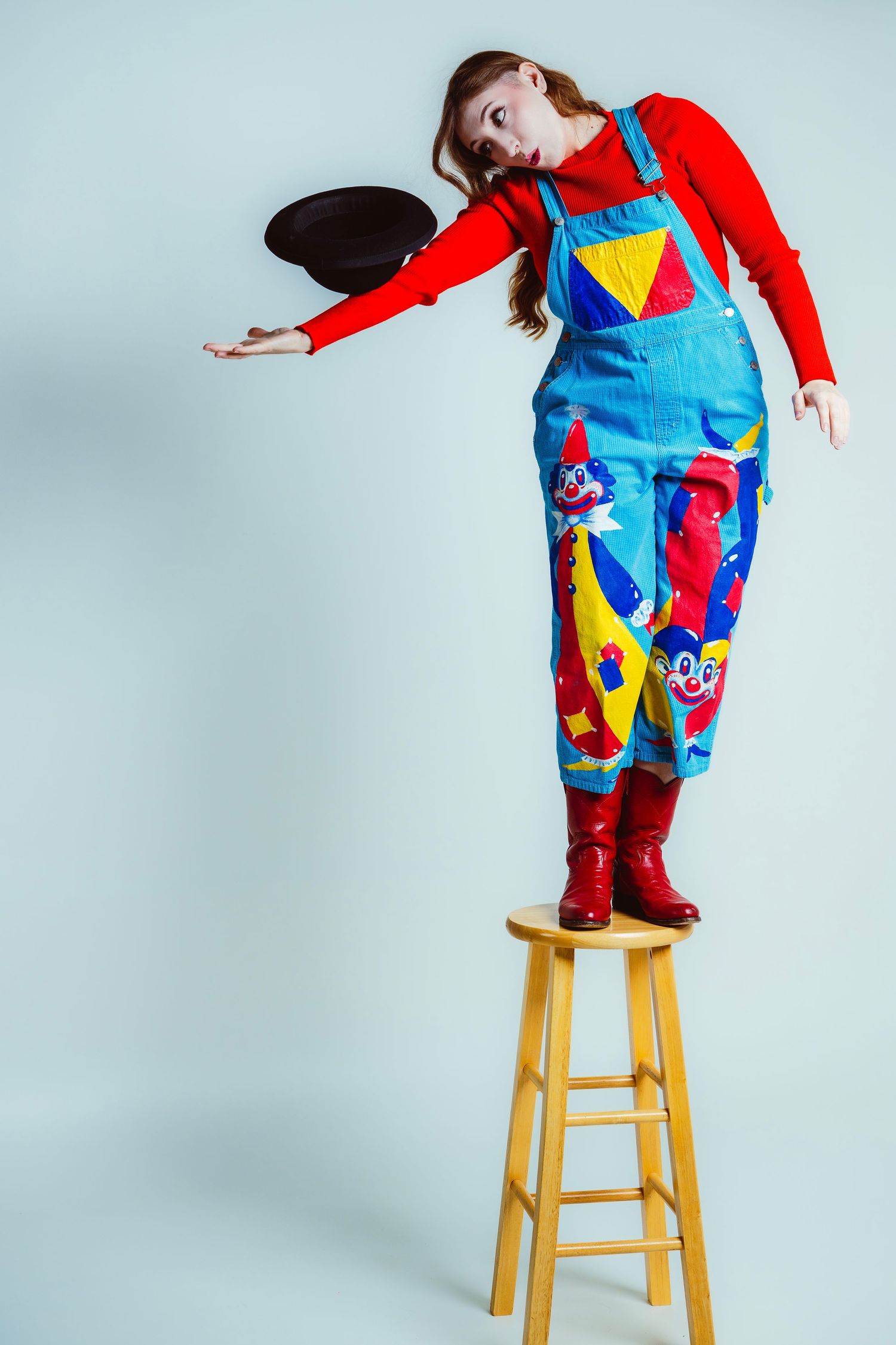 Person in bright overalls performs balancing acts on wooden stool against light background.