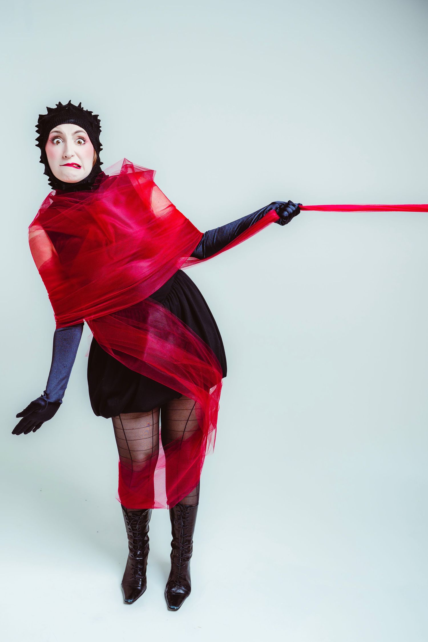 Figure in dramatic red flowing fabric and black boots poses against light background while being pulled by red ribbon.