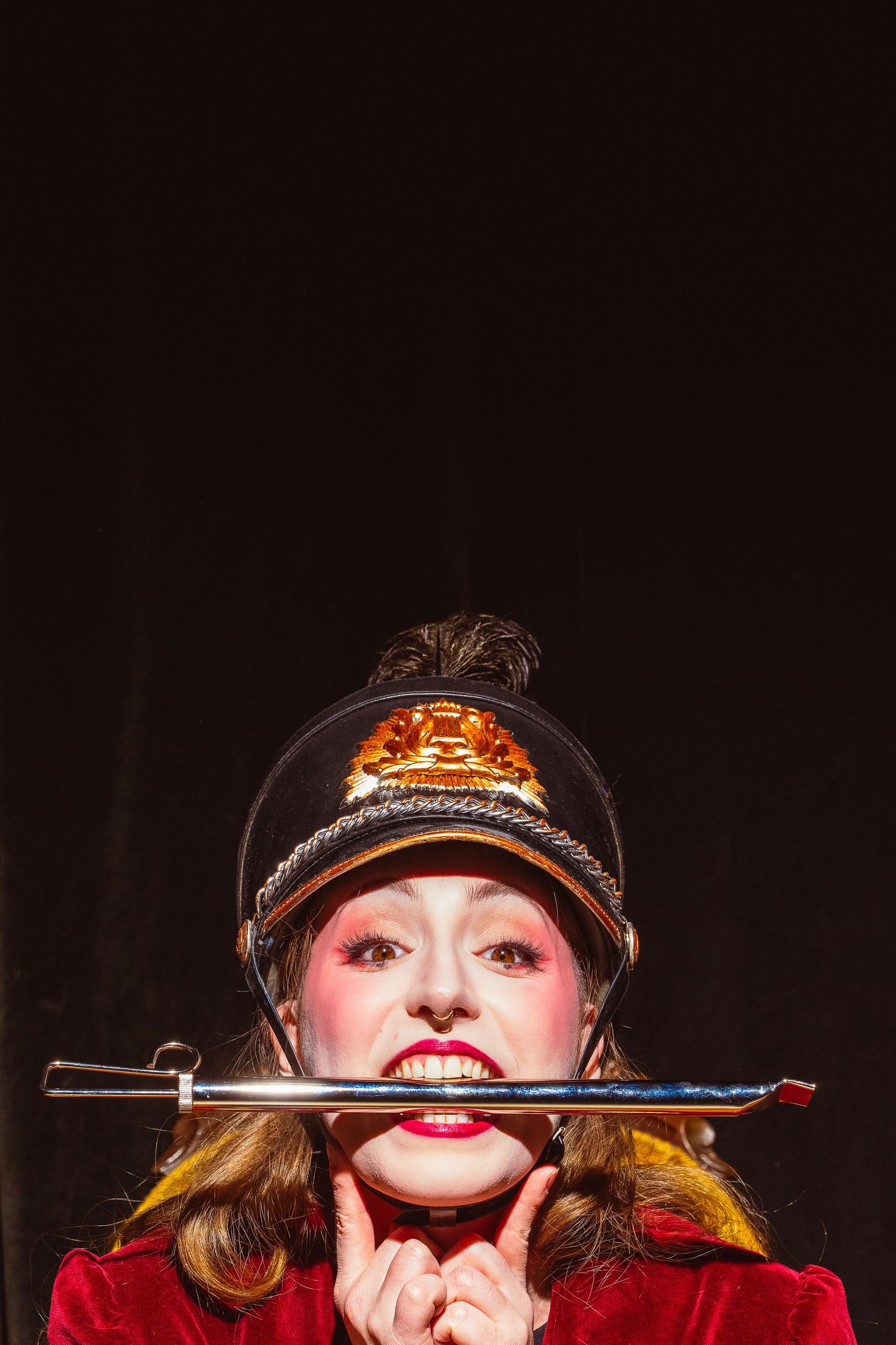 A performer in a black cap and red outfit holds a sword horizontally in a dramatic dark-lit stage pose.