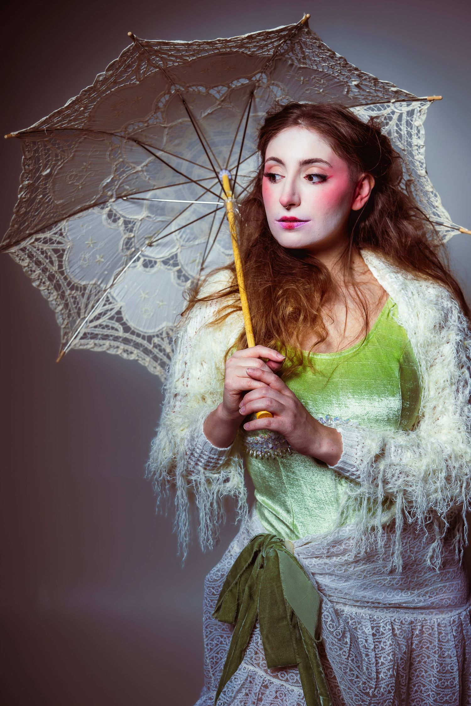 Figure in a lime green dress holds delicate white lace parasol with dramatic theatrical makeup and flowing hair.