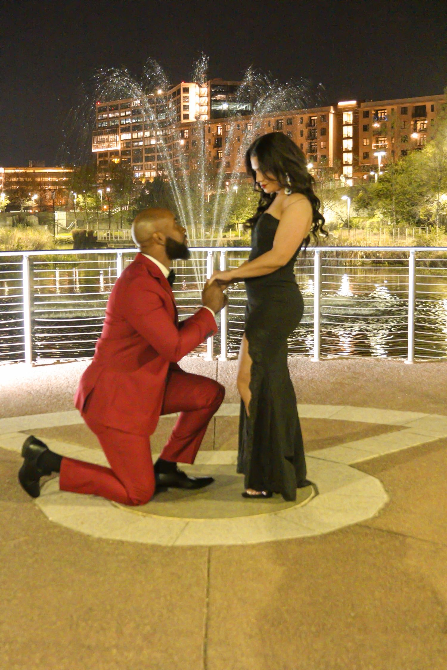 A romantic proposal scene at night by a fountain with illuminated buildings and water features in the background.