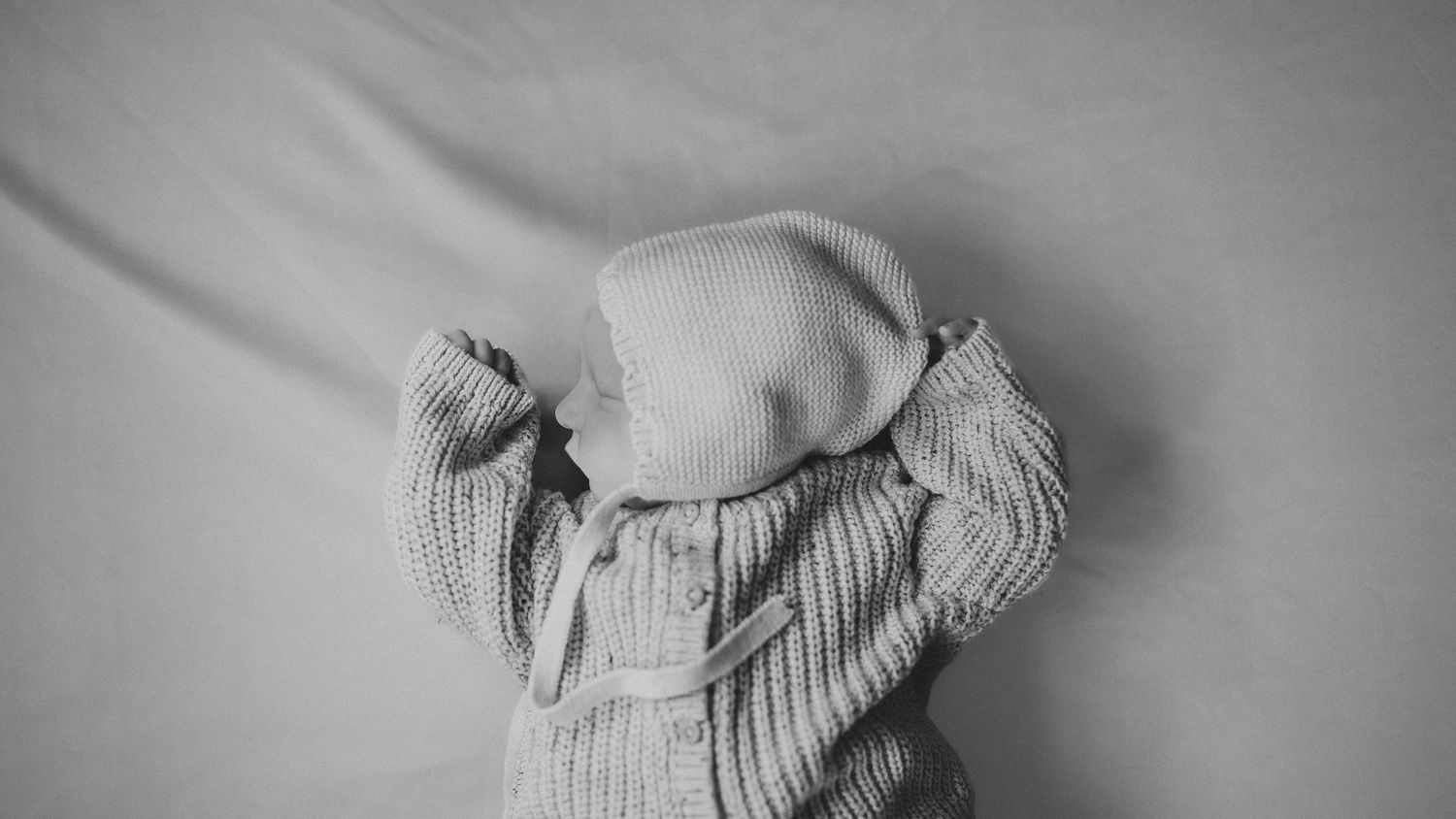 Black and white photo of a newborn baby lying face down in a knit sweater and hat on a white blanket.