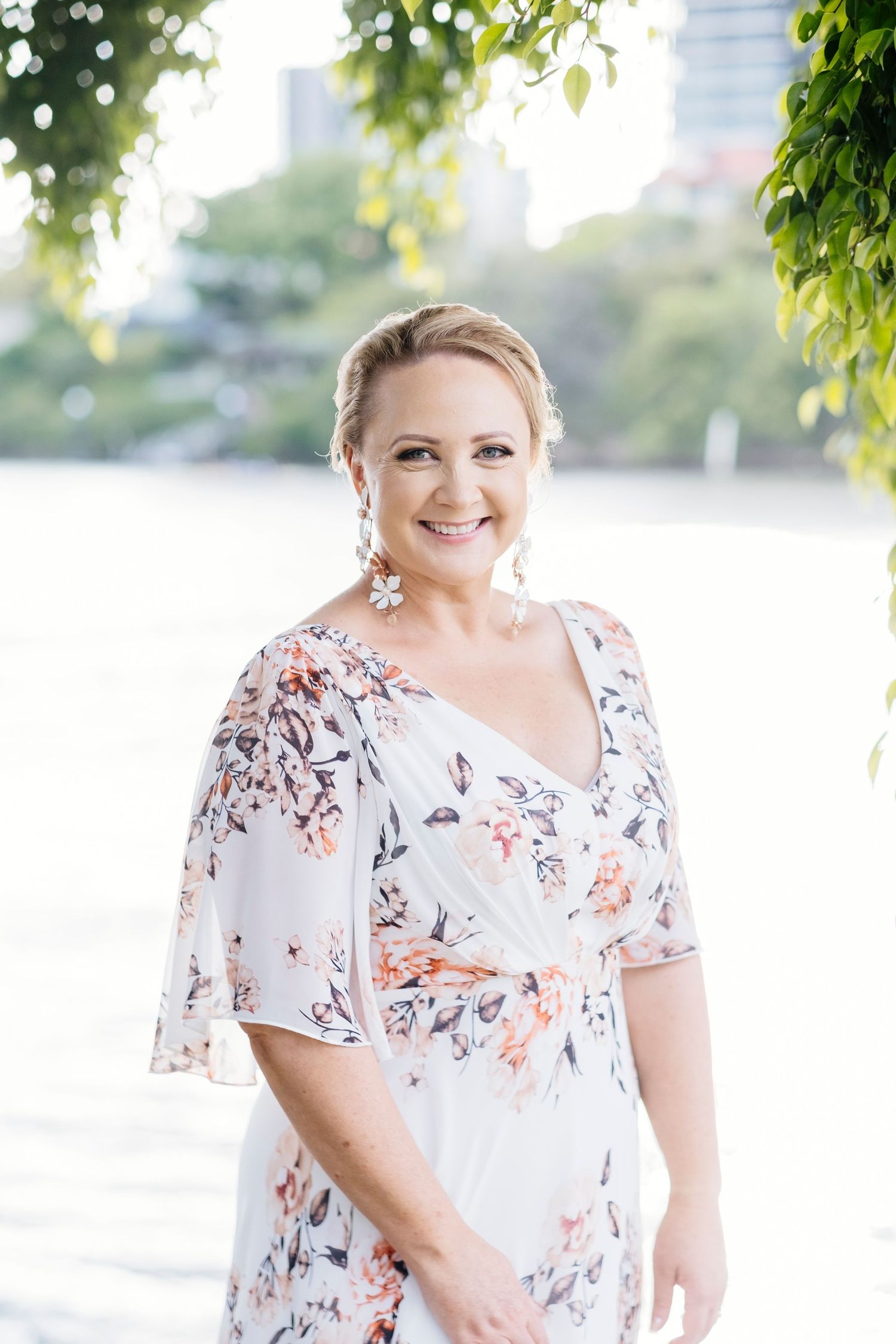 A woman in a white floral dress with flutter sleeves poses outdoors with greenery and city buildings in the background.