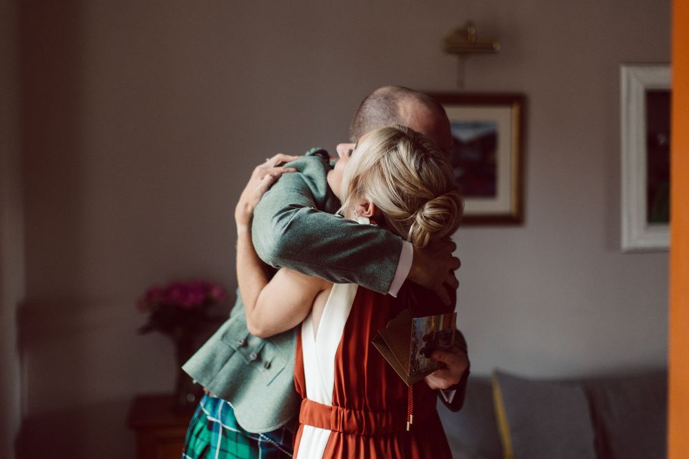 Two people share an emotional embrace in a dimly lit room with warm lighting and framed artwork on the wall.