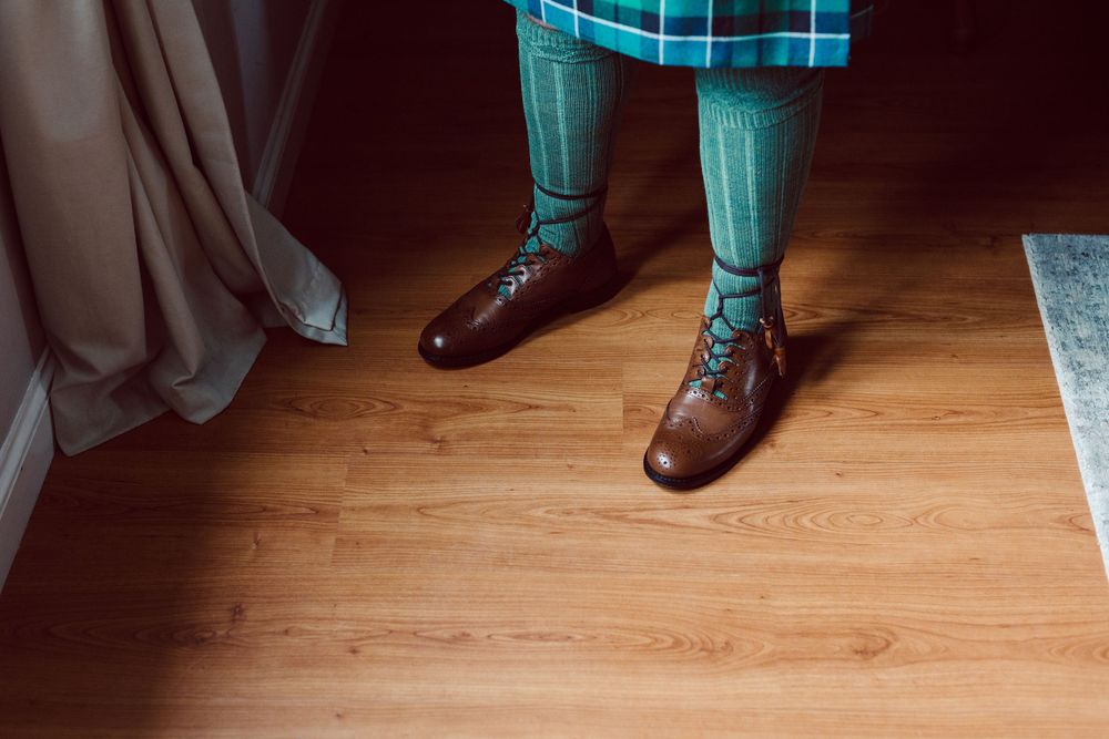 Brown leather dress shoes paired with turquoise plaid socks on hardwood flooring.