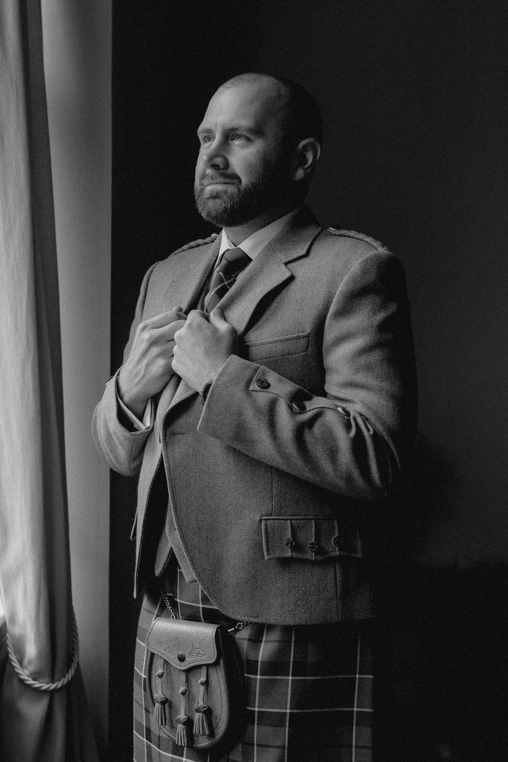 A groom in a formal gray suit jacket and traditional tartan kilt stands by a window in dramatic black and white lighting.