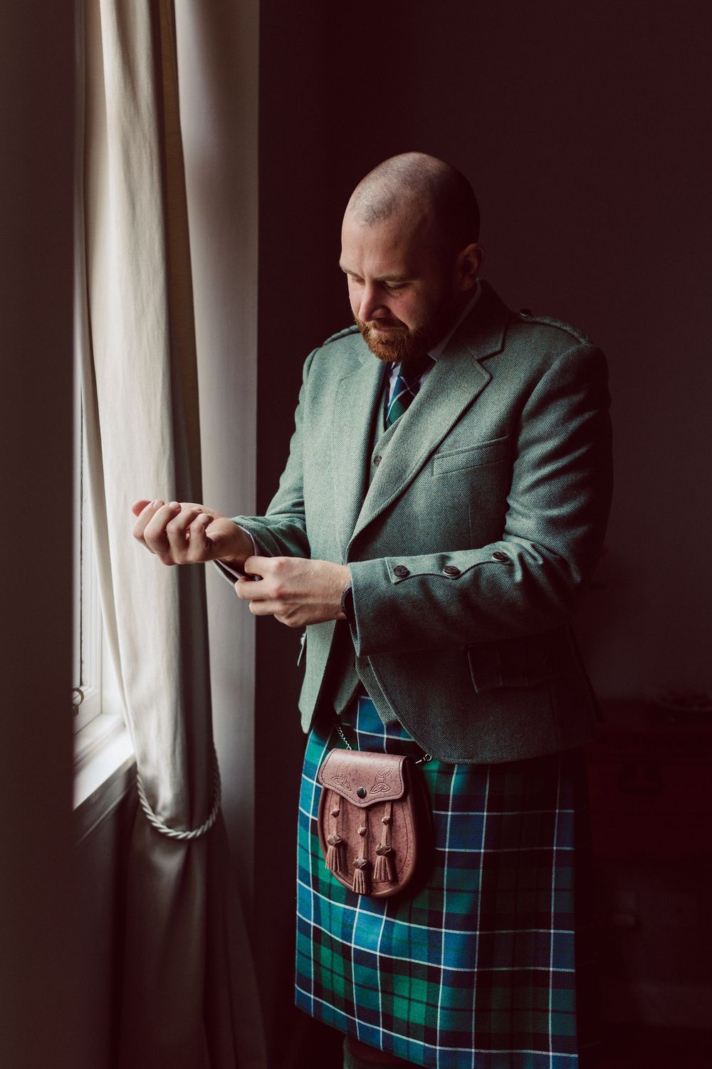 A person in a green jacket and tartan kilt adjusts their outfit near a window in moody lighting.
