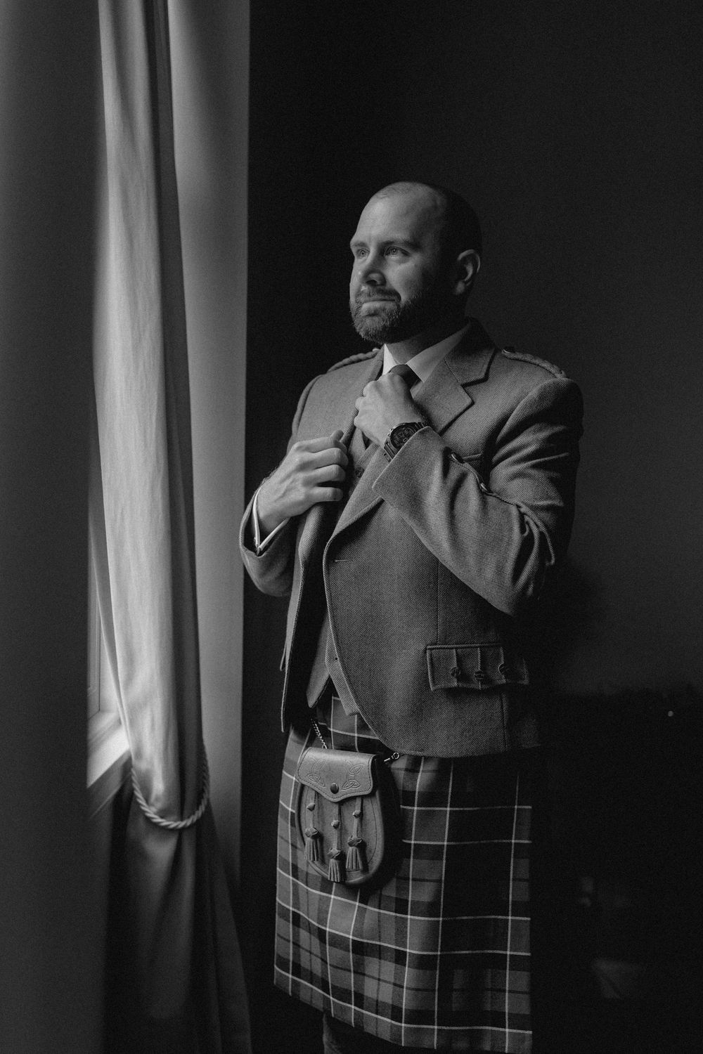 A black and white photo series shows someone wearing a suit and plaid kilt while adjusting their tie in dramatic lighting.