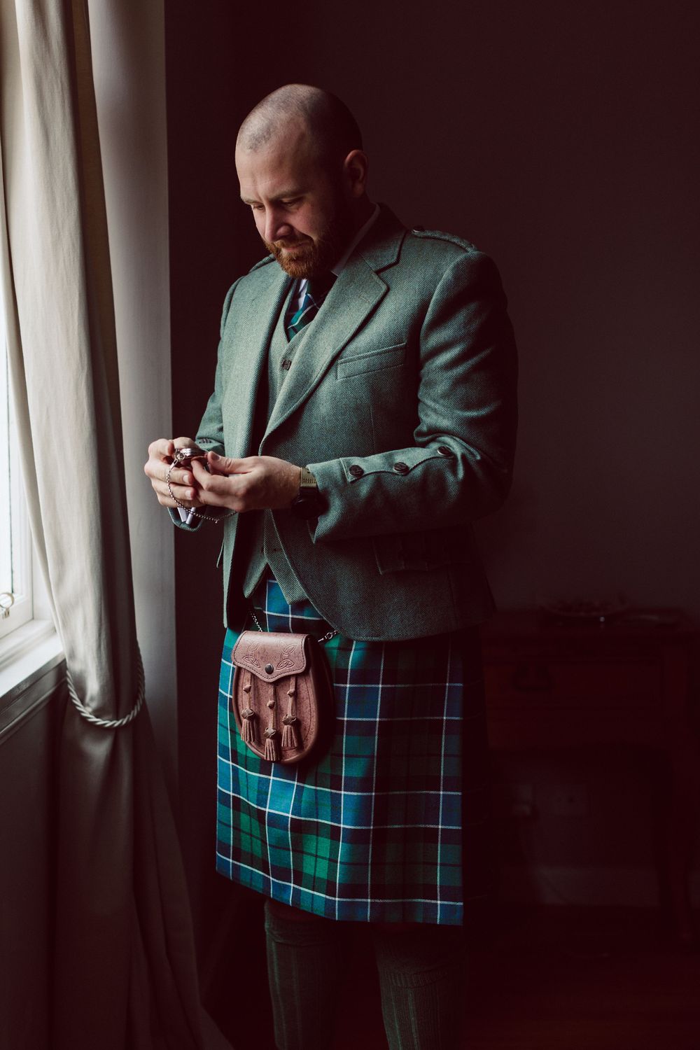Person wearing a green blazer and blue-green tartan kilt stands by window while checking their phone in moody lighting.