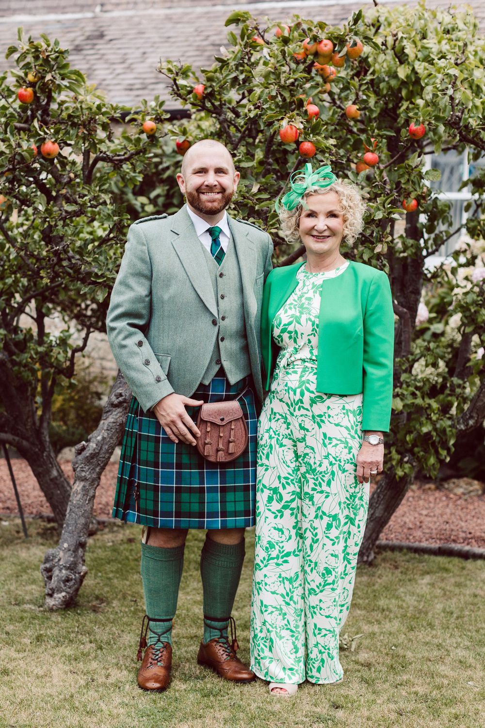 A couple poses outdoors in coordinating green attire with a Scottish kilt and floral dress against an apple tree backdrop.