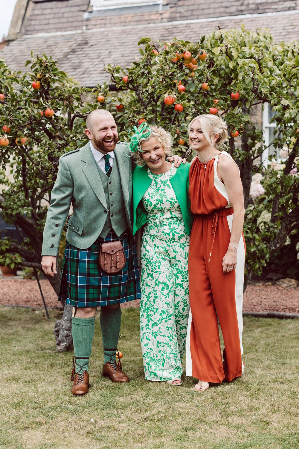 A wedding party poses together in a garden with green and floral outfits against a brick wall backdrop.