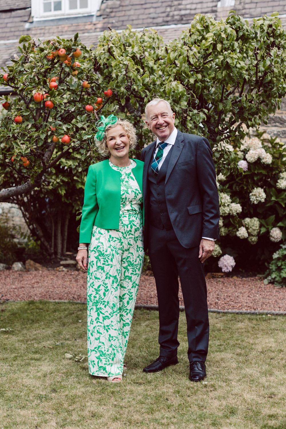 A couple stands together in a garden with blooming orange flowers and white roses in the background.