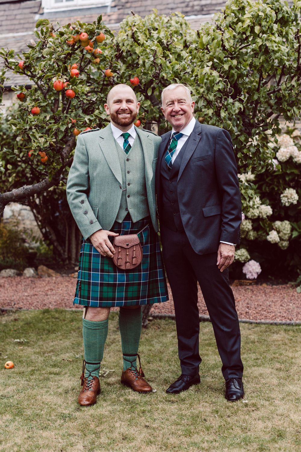 People in traditional Scottish formal wear pose together in a garden with flowering bushes.