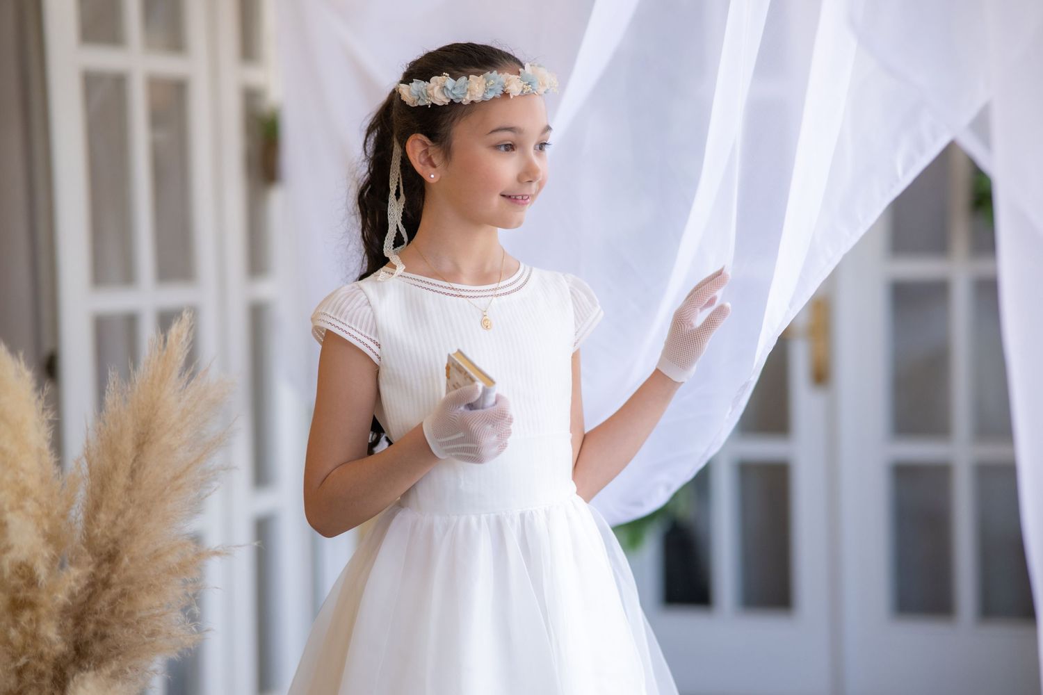 A person in a white dress stands by sheer curtains and pampas grass in a light-filled room.