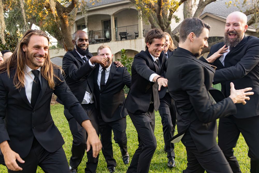 A wedding party in black suits jumps together on a lawn for a fun group photo.