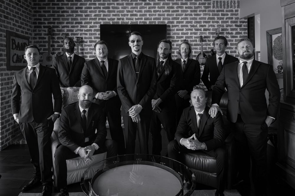 A black and white photo shows a group of men in suits standing against a brick wall.